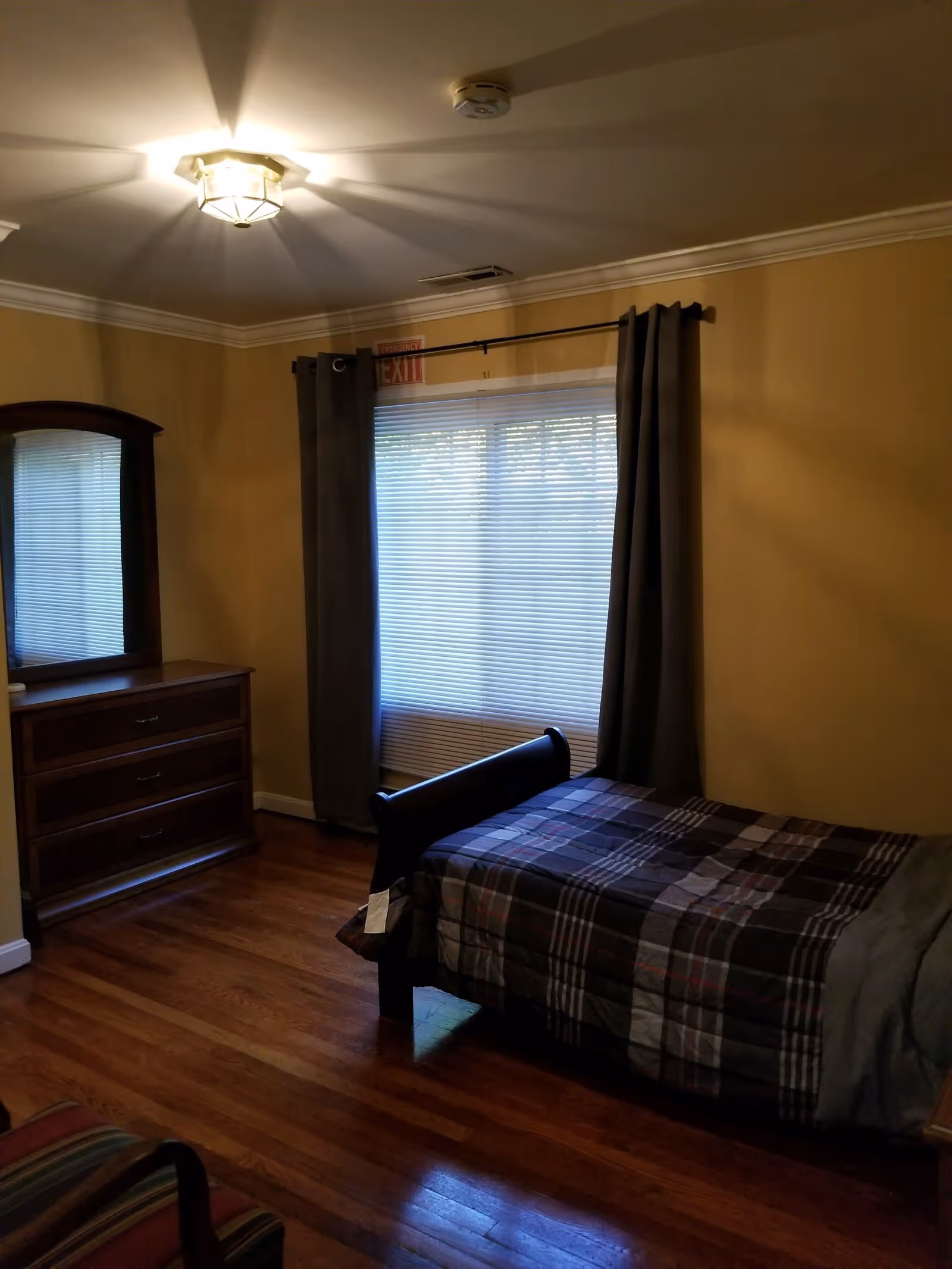 A bedroom with wooden flooring, a single bed with a plaid blanket, a wooden dresser with a mirror, a window with closed blinds and dark curtains, and a ceiling light fixture.