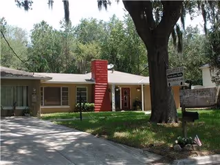 Single-story residential building with a red brick chimney, beige exterior walls, and a front yard with green grass and large trees. A curved driveway leads to the entrance, and a street sign is visible near the tree in the foreground.