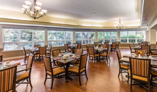Spacious dining room with multiple set tables, chandeliers, and large windows overlooking trees.