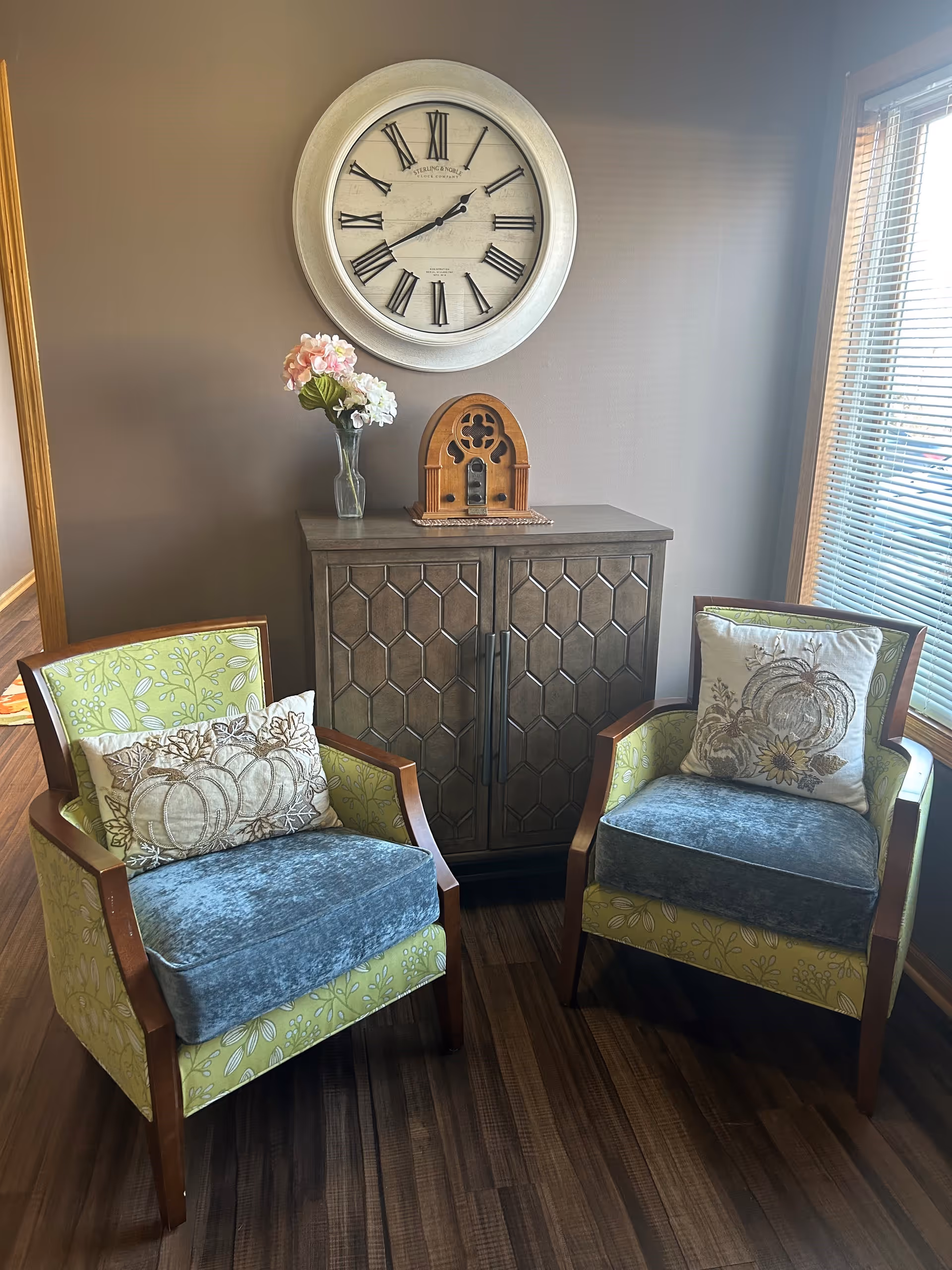 Two upholstered armchairs with green floral fabric and blue cushions, each with a decorative pillow, are positioned on either side of a wooden cabinet with a geometric pattern. On top of the cabinet is a vase with pink and white flowers and a vintage-style wooden radio. Above the cabinet hangs a large round wall clock with Roman numerals. A window with blinds is visible to the right, letting in natural light.