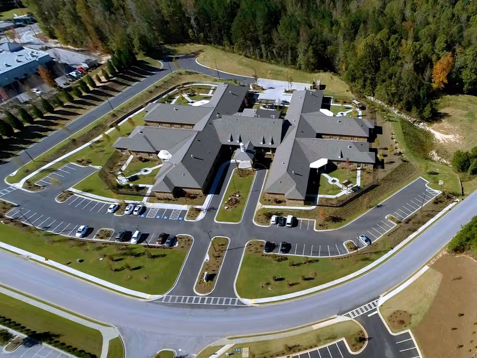 Aerial view of a large memory care facility named Hope Center Memory Care, showing multiple connected buildings with gray roofs, surrounded by parking lots, landscaped green areas, and roads. The facility is bordered by trees and open land.