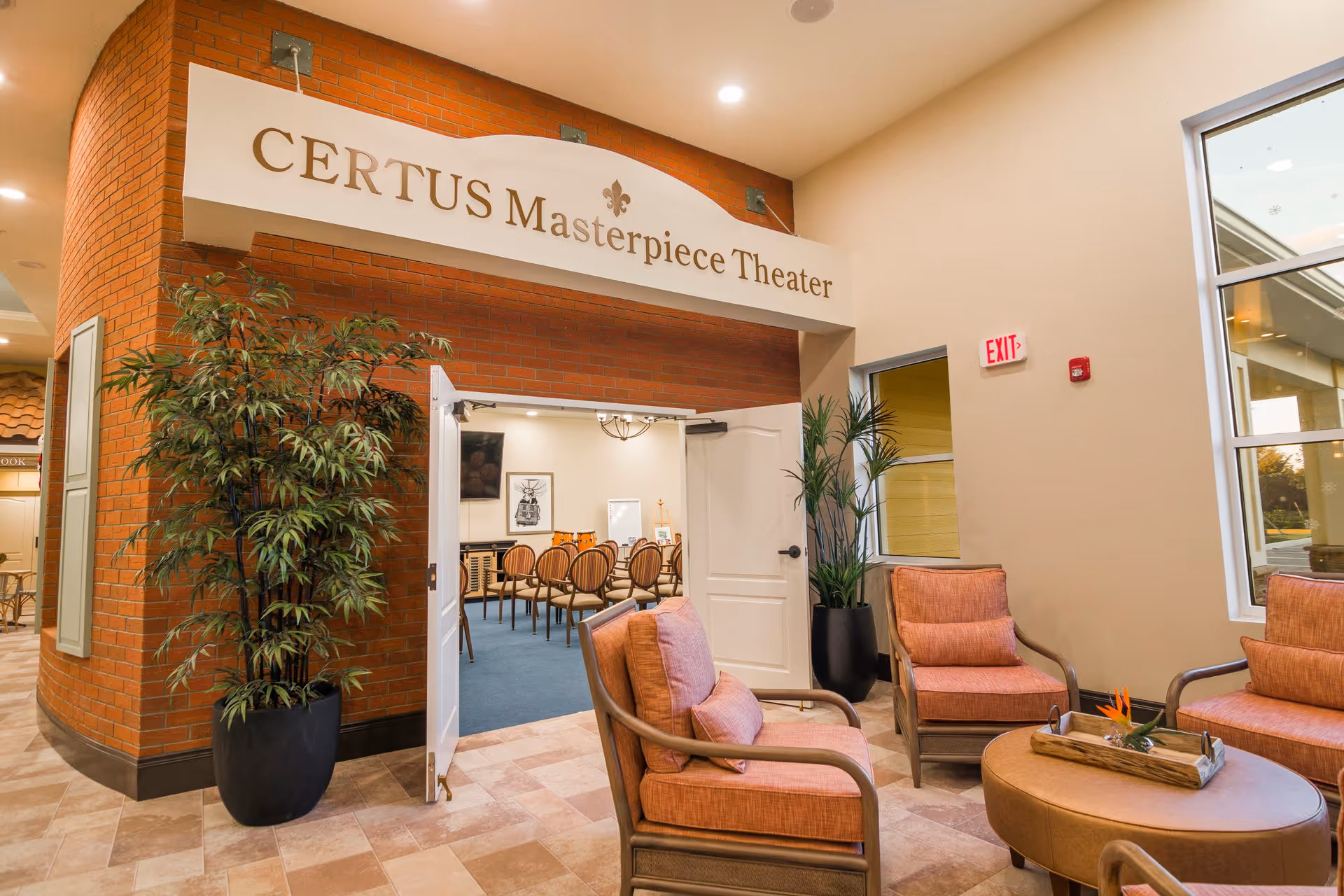 Interior view of CERTUS Premier Memory Care Living showing the entrance to the CERTUS Masterpiece Theater with open double doors. The area outside the theater has comfortable seating with cushioned chairs and a round ottoman table with a decorative tray. There are large potted plants on either side of the theater entrance and a window letting in natural light.