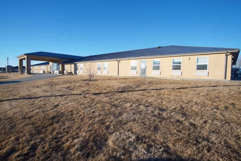 Exterior view of a single-story assisted living residence building with a covered entrance, beige walls, multiple windows, and a dry grassy lawn under a clear blue sky.