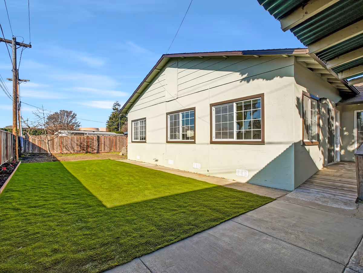 Outdoor view of a senior living facility showing a green lawn, concrete pathway, and the exterior wall of a building with three windows under a clear blue sky.
