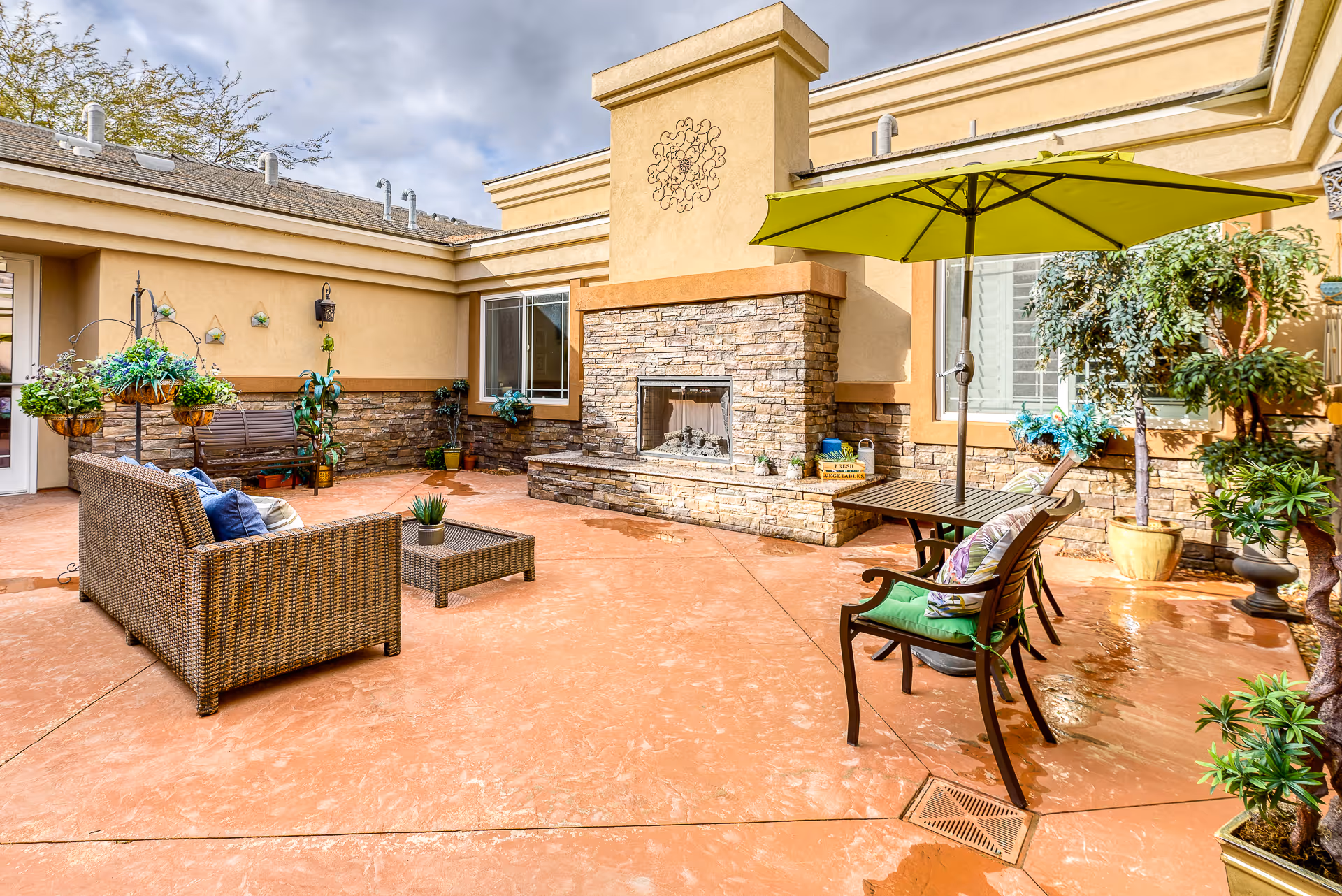 Sunny courtyard patio with wicker seating, a stone fireplace, potted plants and a green umbrella.