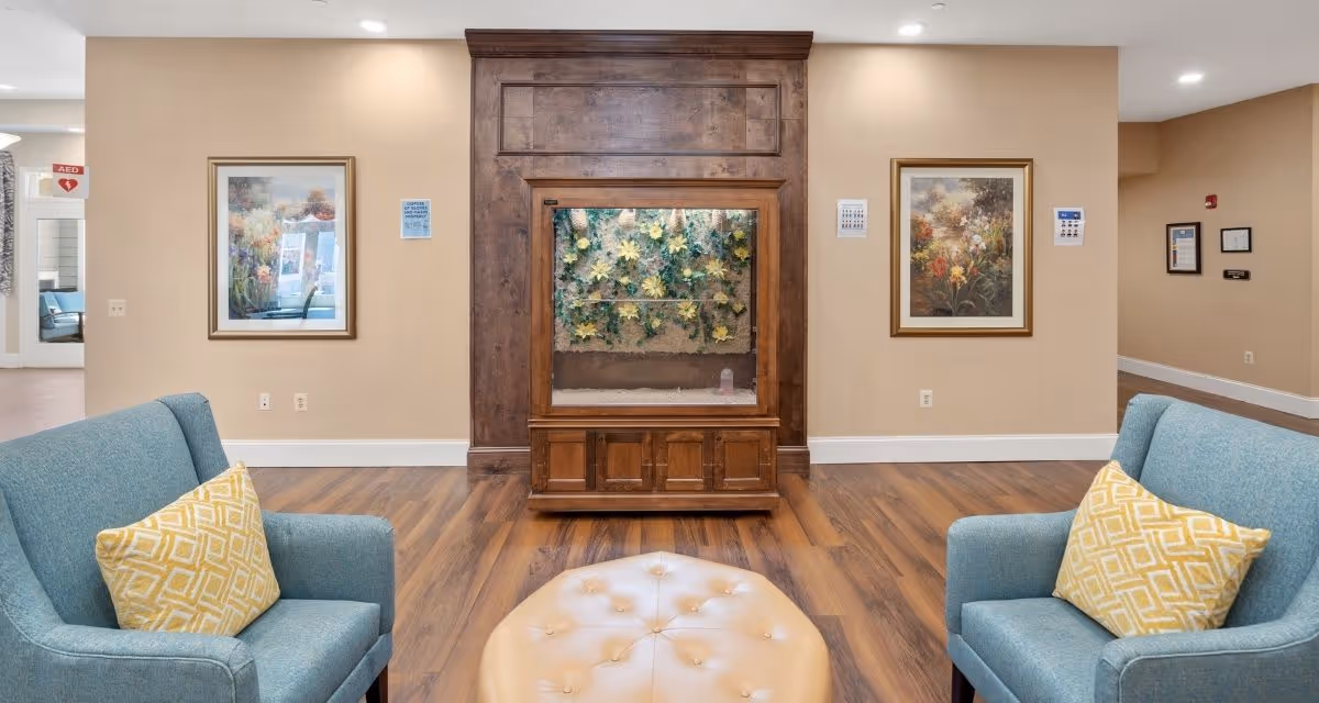 A cozy seating area in a senior living facility with two blue armchairs, each with a yellow patterned pillow, facing a wooden cabinet with a glass display filled with yellow flowers. The room has wood flooring, beige walls, and framed floral artwork on the walls.