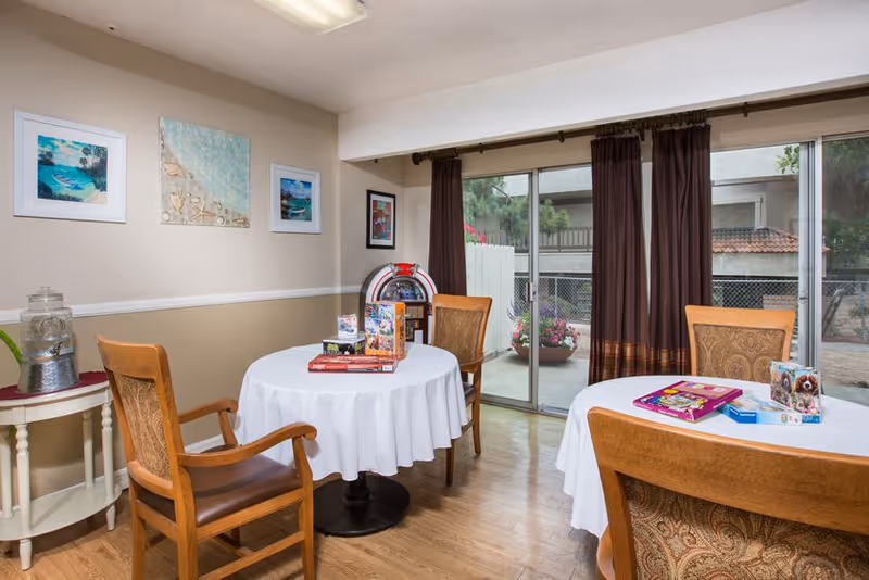 Communal dining/activity room with round tables draped in white tablecloths, wooden chairs, wall art, and sliding glass doors to a patio.