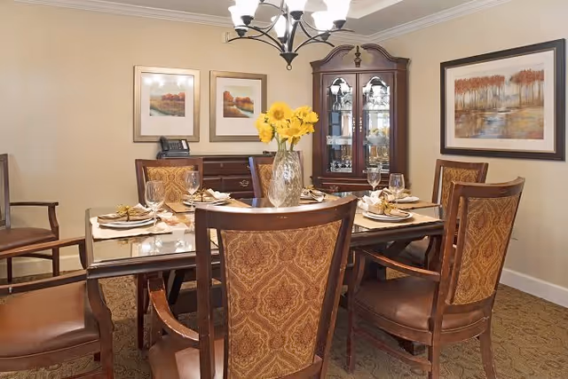 A formal dining room with a rectangular glass-top table set for six people. The table has elegant place settings with plates, napkins, and wine glasses. A vase with yellow flowers is centered on the table. The room features upholstered wooden chairs with patterned fabric backs and seats. On the walls are framed landscape paintings, and a wooden china cabinet with glass doors is visible in the background. A chandelier with multiple lights hangs above the table.