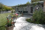 Small courtyard with a paved walkway, benches, potted flowers, lawn, and a shaded patio area with chairs and climbing vines.