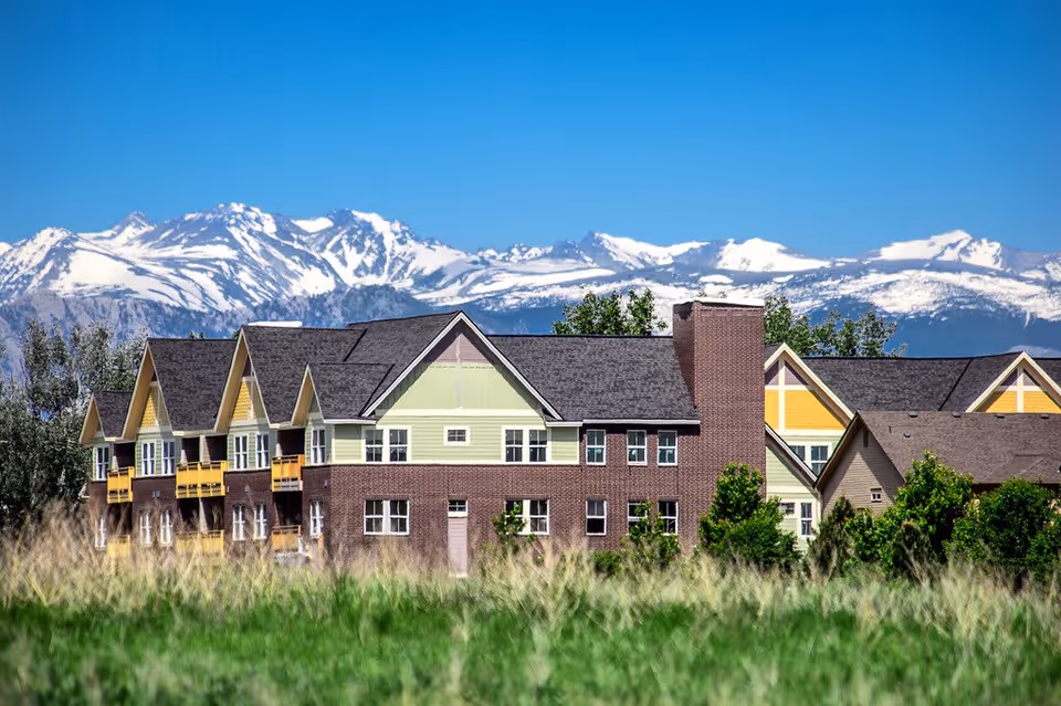 Exterior view of a multi-story residential building with a brick lower facade and light green and yellow upper sections, set against a backdrop of snow-capped mountains and a clear blue sky, with green grass and trees in the foreground.