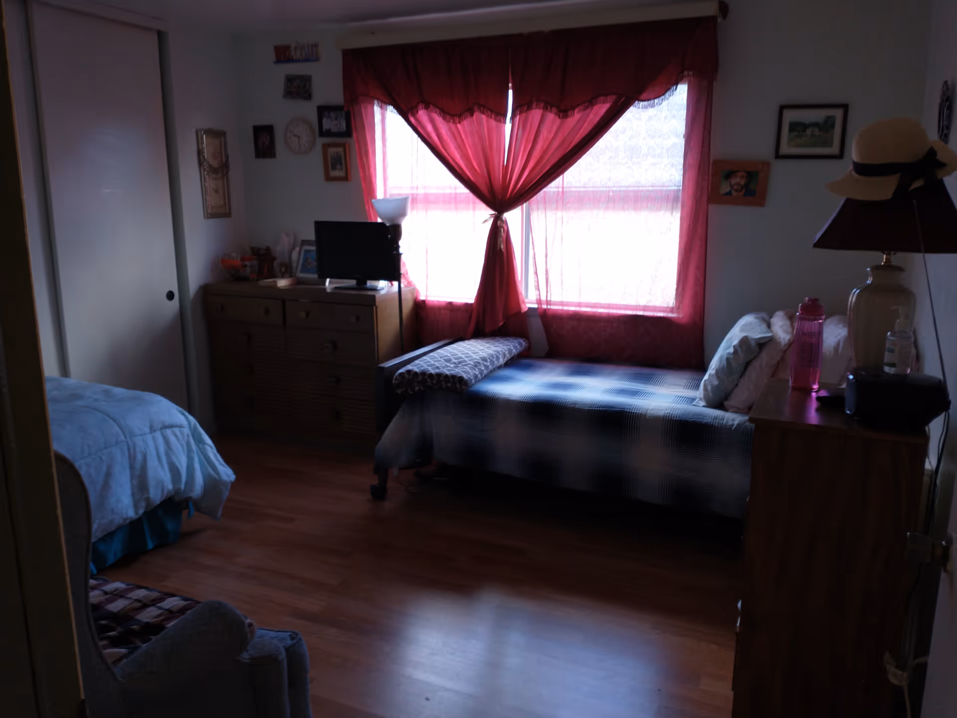 A dimly lit bedroom with a single bed covered in a blue and white checkered blanket, a wooden dresser with a small TV on top, a window with red curtains letting in some light, and a nightstand with a lamp, water bottle, and other small items.