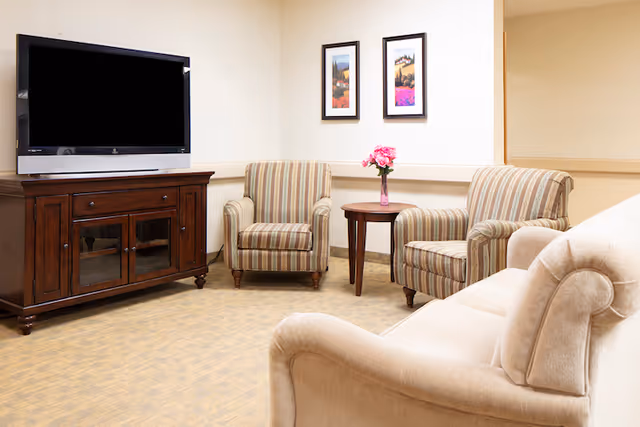 Small sitting room with a couch, two striped armchairs, a round side table with flowers, a wooden TV console and a wall-mounted flat-screen TV.