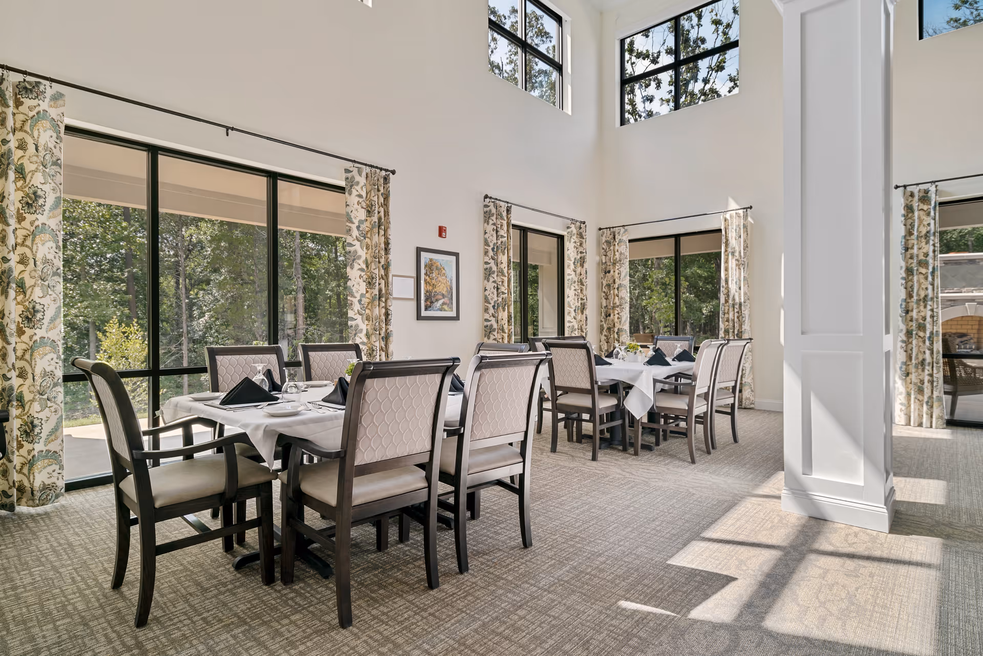 A bright dining room with two tables set for meals, each surrounded by eight cushioned chairs. Large windows with floral curtains let in natural light and offer views of green trees outside. The room has high ceilings and a carpeted floor.