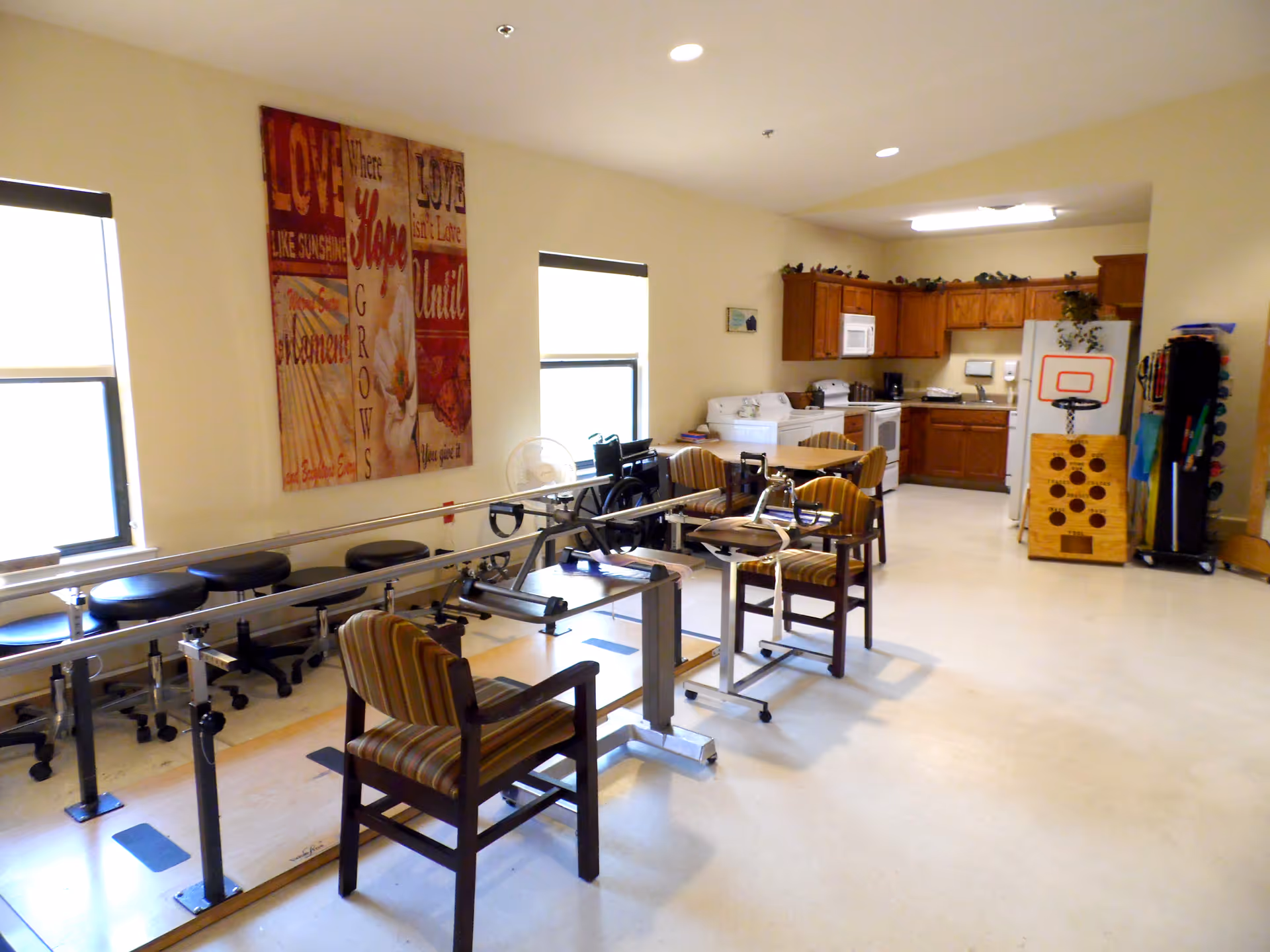 A rehabilitation room with parallel bars for walking exercises, several striped chairs, stools along the wall, and a kitchenette area with wooden cabinets, a microwave, stove, and refrigerator. The room has two windows and motivational wall art.