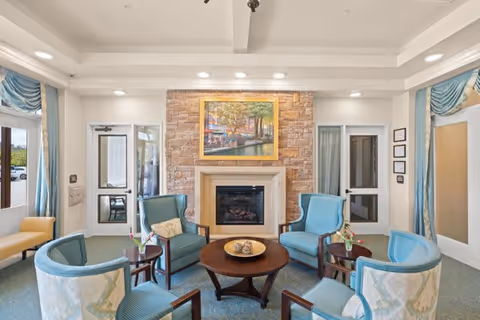 A cozy seating area in a memory care community featuring six blue and beige upholstered chairs arranged around a round wooden coffee table with decorative items. The room has a stone fireplace with a framed painting above it, light blue curtains, and doors with glass panels on either side of the fireplace.