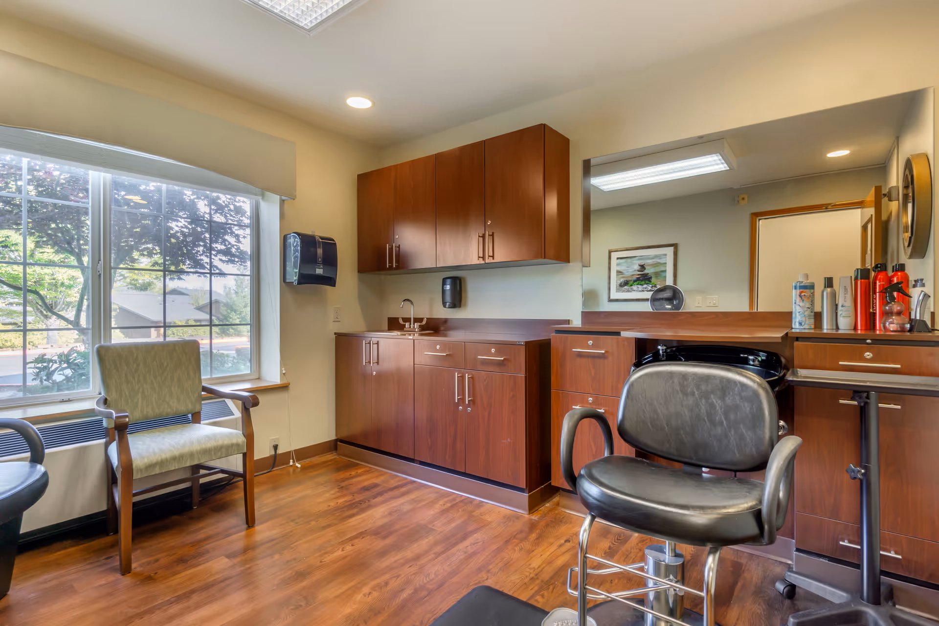 A salon-style interior with a barber chair, wooden cabinets, countertop sink and a large window overlooking trees.