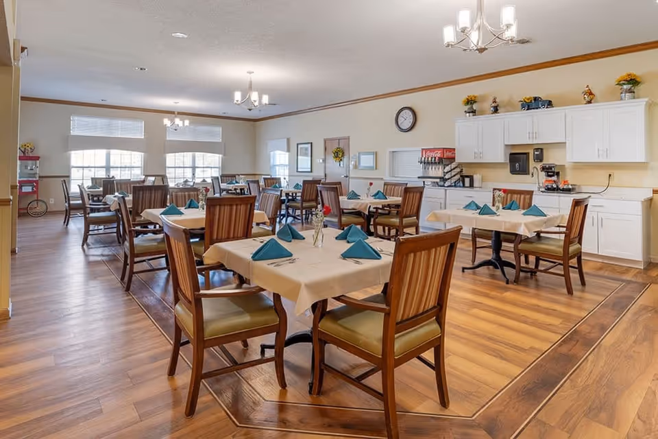 A bright dining room with several tables covered with beige tablecloths and green folded napkins. Each table has wooden chairs with green cushions. The room has large windows letting in natural light, wooden flooring, and white cabinets along one wall with a beverage dispenser and coffee machine. Ceiling lights provide additional illumination.