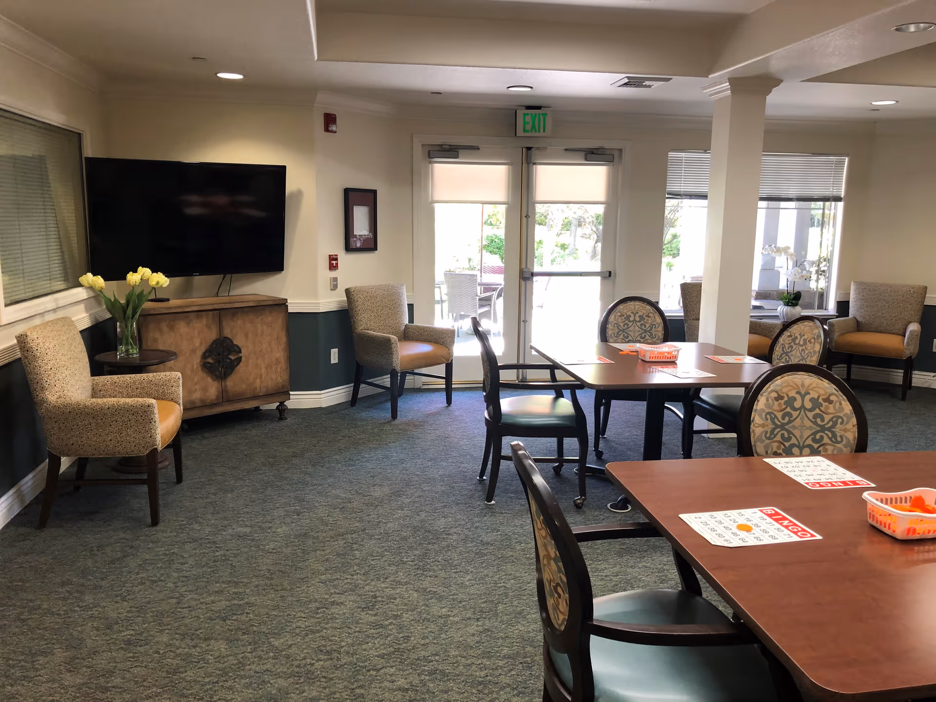 A common area in a senior living facility with several chairs arranged around tables. Bingo cards and orange markers are on the tables. There is a TV mounted on the wall above a wooden cabinet, a vase with yellow flowers on a small table, and glass doors leading outside.