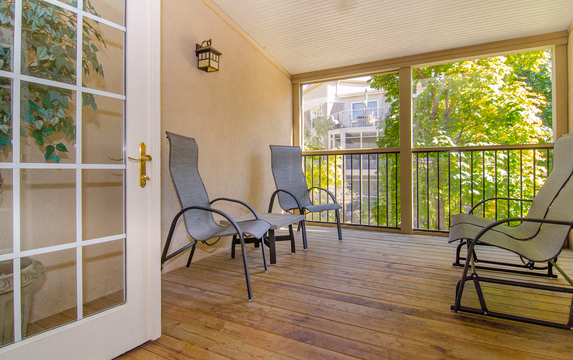 Covered wooden balcony with several lounge chairs overlooking trees and a neighboring building.
