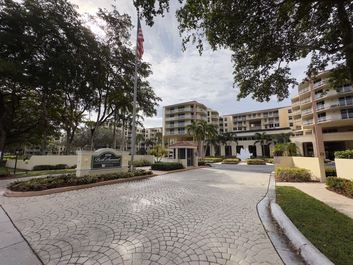 Entrance and driveway of the Park Summit senior living complex with a flagpole, sign, fountain and multi-story buildings.