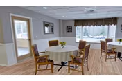 A bright dining area with round tables covered in white tablecloths, each surrounded by wooden chairs with patterned cushions. Large windows with valance curtains allow natural light to fill the room, and a door with glass panels leads to another area. The floor is wood-patterned, and the walls are painted light gray with white wainscoting.