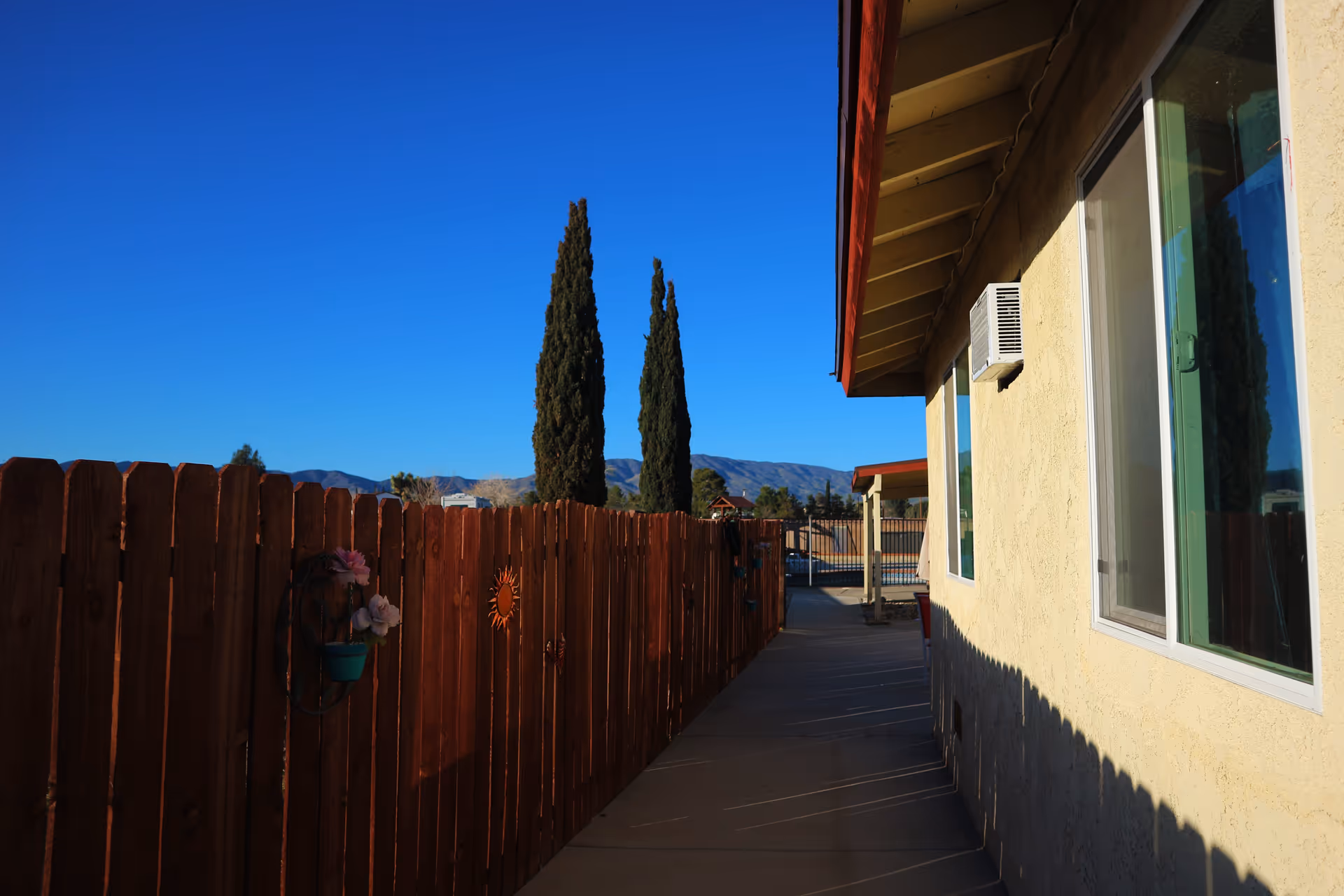 Outdoor walkway beside a yellow building with windows and an air conditioning unit, bordered by a wooden fence decorated with flower pots and sun ornaments, with tall trees and mountains visible in the background under a clear blue sky.