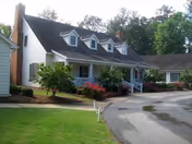 Exterior view of a single-story assisted living facility building with white siding, a dark roof with dormer windows, a front porch with white railings, and landscaped bushes and flowers along the walkway and driveway.