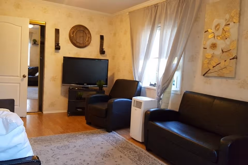 Cozy living room with black leather sofa and armchair, a TV on a stand, sheer-curtained window, and wall decorations.