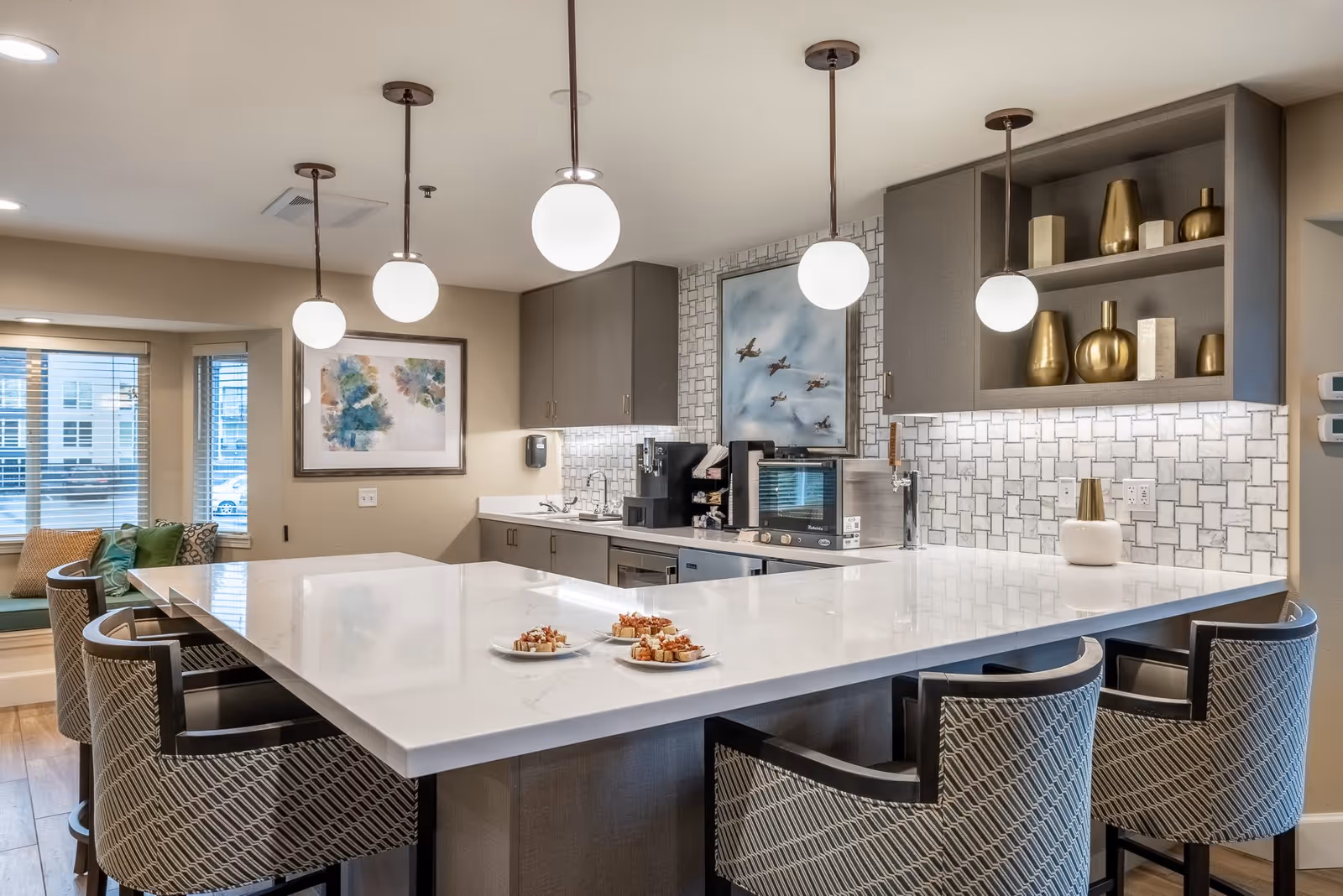 Modern kitchen area with a large white marble island countertop surrounded by patterned cushioned chairs. The kitchen features gray cabinets, a tiled backsplash, and pendant lights hanging from the ceiling. There are decorative gold vases on open shelves and framed artwork on the walls. Plates with small food servings are placed on the island.