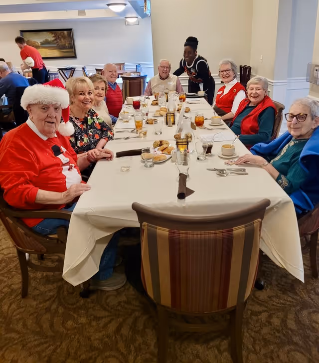 A group of elderly people sitting around a long dining table in a senior living facility, enjoying a meal together. One person is wearing a Santa hat and festive sweater, suggesting a holiday celebration. A staff member is standing and smiling behind the group. The table is set with plates, glasses, and various food items.