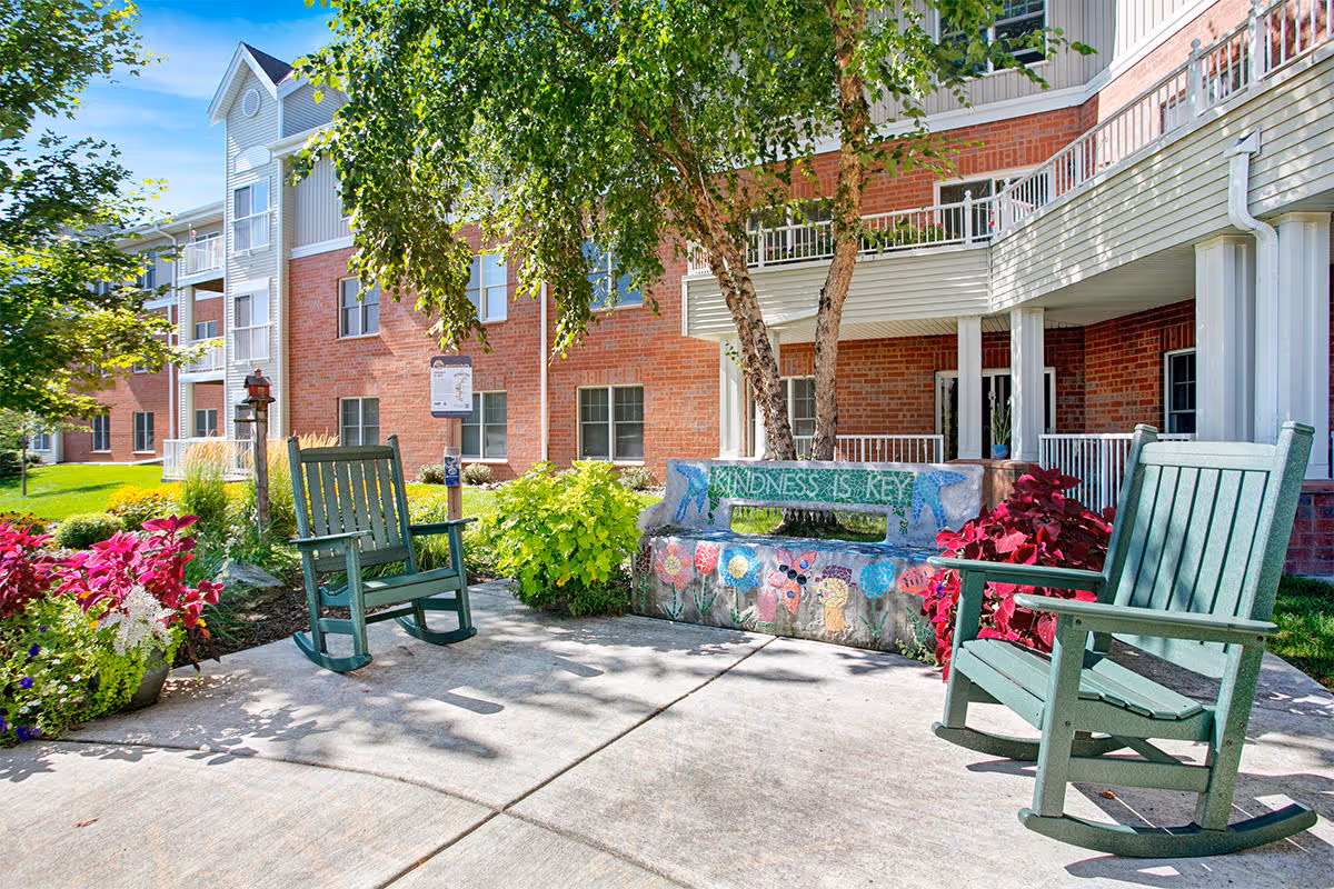 Outdoor courtyard of a brick senior living building with green rocking chairs, a decorated bench reading "Kindness is Key", plants and balconies.