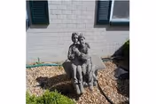 Stone sculpture of two seated figures in a small landscaped area in front of a light brick building with shuttered windows.