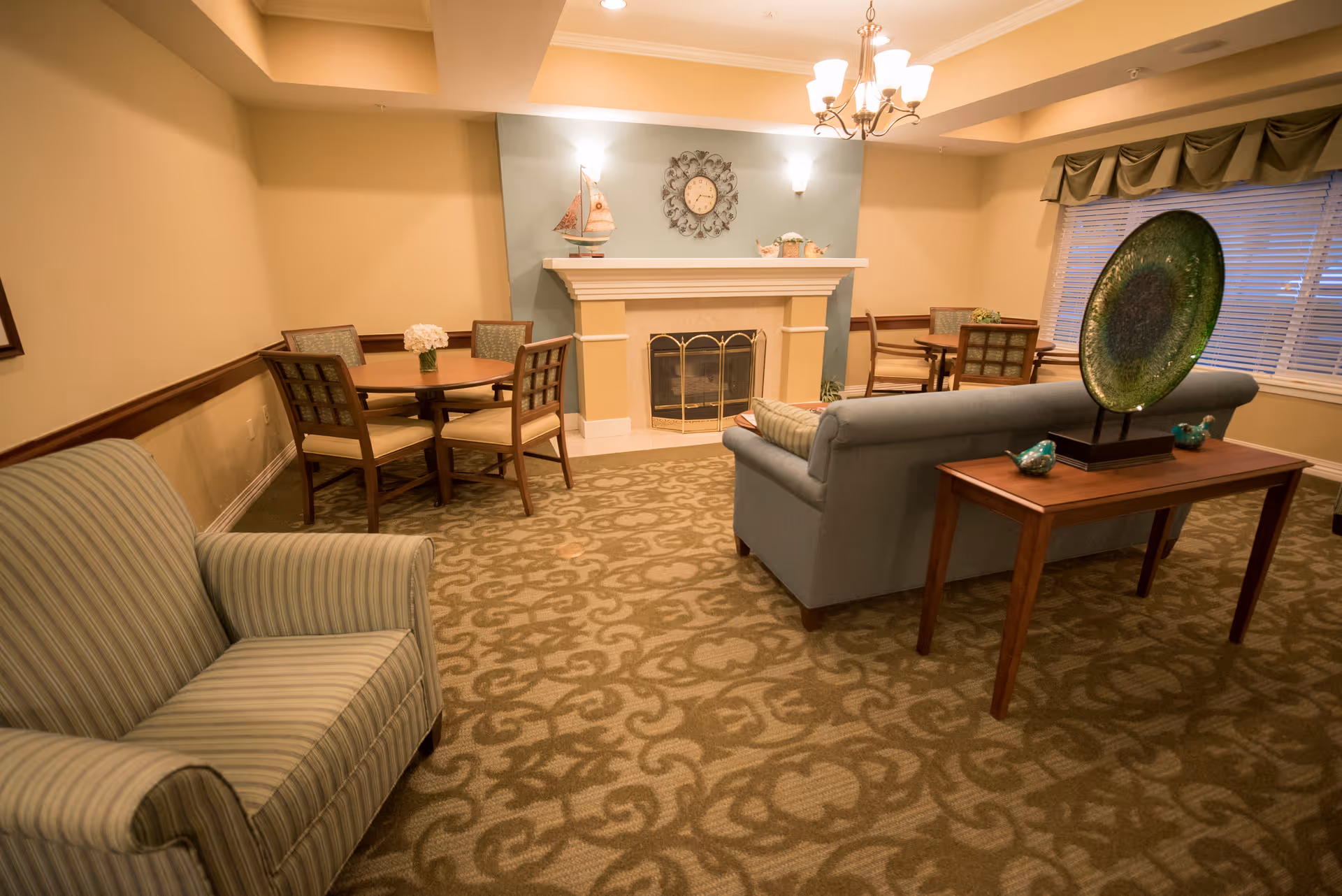 Cozy common living room in a senior facility with a sofa and armchairs arranged around a fireplace and small dining tables.