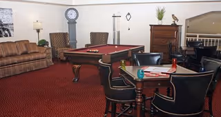 A cozy common area with a red carpet featuring a pool table in the center, surrounded by a brown striped sofa, two armchairs, a tall grandfather clock, a wooden chest of drawers with a decorative bird and plant on top, and a round table with four black leather chairs. The walls are light-colored and decorated with a framed picture and a wall-mounted coat rack.