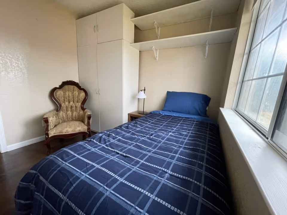 A small bedroom with a single bed covered in a blue plaid blanket and a matching blue pillow. Next to the bed is a wooden nightstand with a small lamp. There is a vintage upholstered armchair with wooden frame in the corner. A white wardrobe with cabinets and two white shelves mounted on the wall are also visible. A large window lets in natural light from the right side.