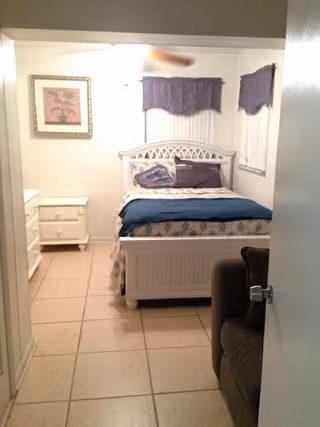 A small bedroom with a white bed frame and a bed made with blue and beige bedding. There are two pillows on the bed, one blue and one beige. A white nightstand with drawers is positioned to the left of the bed. Two windows with purple valances are on the wall behind the bed. A framed picture hangs on the left wall. The floor is tiled, and part of a brown armchair is visible on the right side near the door.