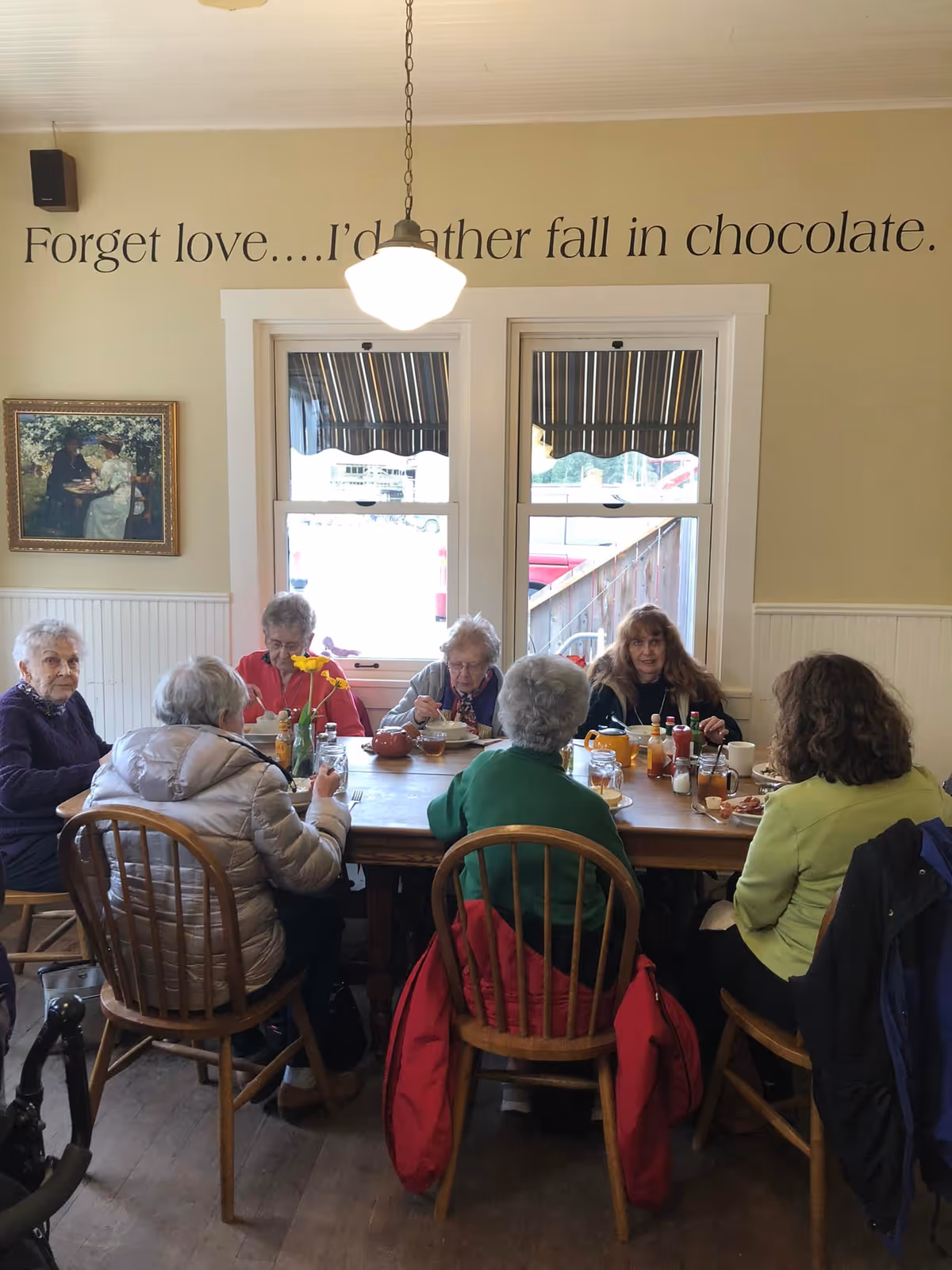 A group of elderly women sitting around a wooden dining table in a cozy room with two windows and a hanging light fixture. On the wall above the windows is a quote that reads, 'Forget love....I'd rather fall in chocolate.' There is a framed painting on the left wall and various condiments on the table.