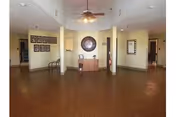 A spacious interior common area with wooden flooring, beige walls, and a ceiling fan. The room features a small wooden table with decorative items and a large circular wall decoration above it. There are framed pictures on the left wall and a mirror on the right wall. Two columns partially divide the space, and doorways lead to other rooms.