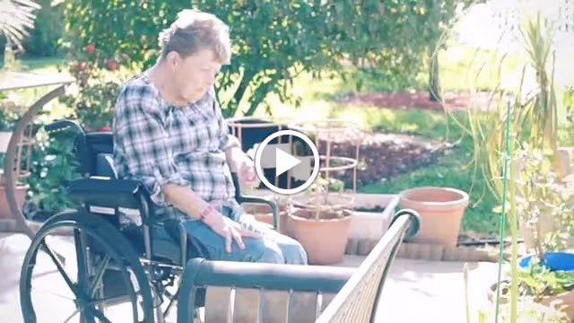 An elderly woman in a wheelchair is sitting outside in a garden area with potted plants and greenery around her. She is wearing a plaid shirt and appears to be looking down at her lap or hands. The scene is bright and sunny.