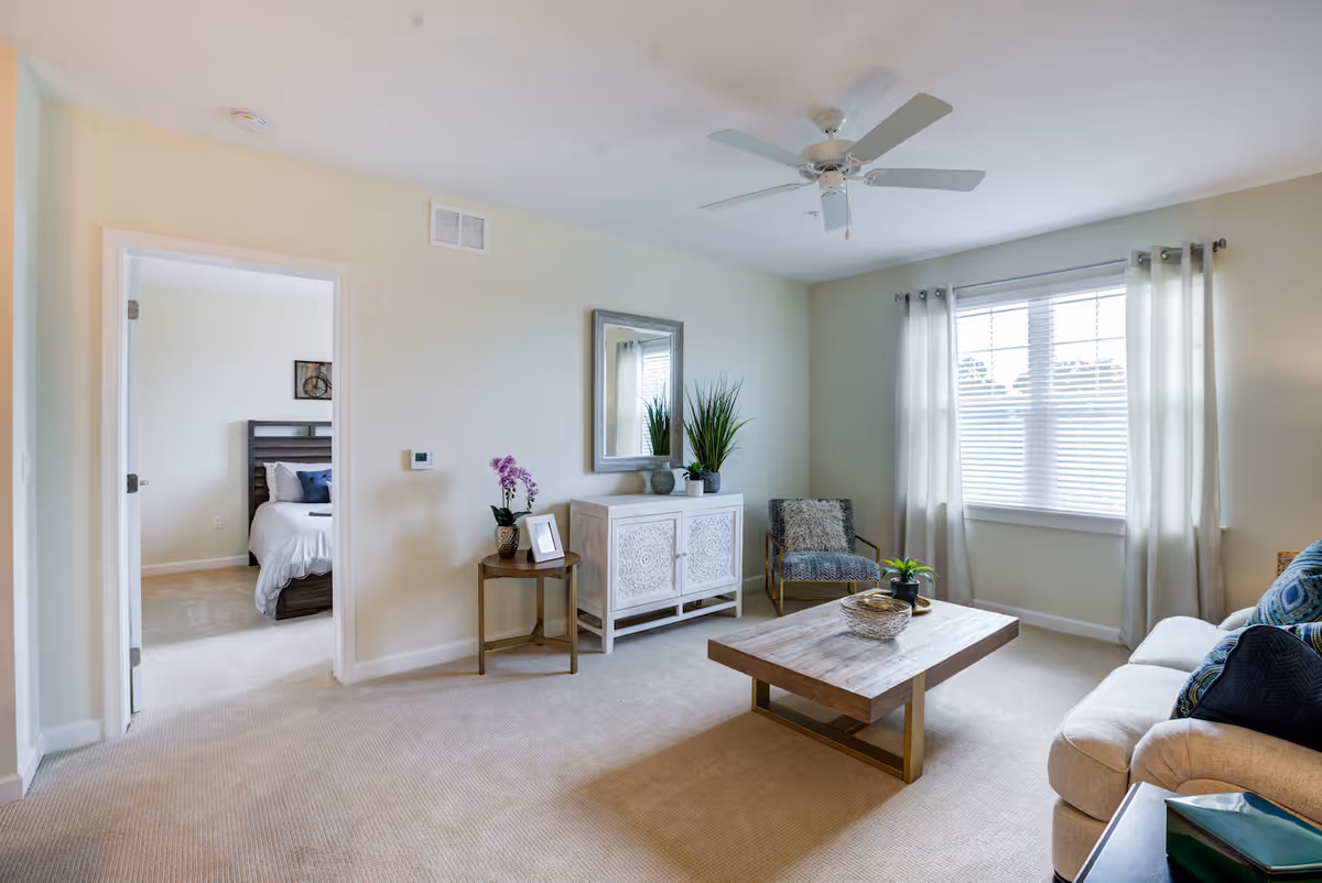 A bright living room with beige carpet and light-colored walls. The room features a beige sofa with blue and patterned cushions, a wooden coffee table with decorative items, a small side table with a purple orchid and a framed photo, a white cabinet with intricate designs topped with plants and a mirror above it, and a single upholstered chair with a furry cushion. A large window with white blinds and light curtains allows natural light to fill the room. An open doorway leads to a bedroom with a bed and wall art visible.