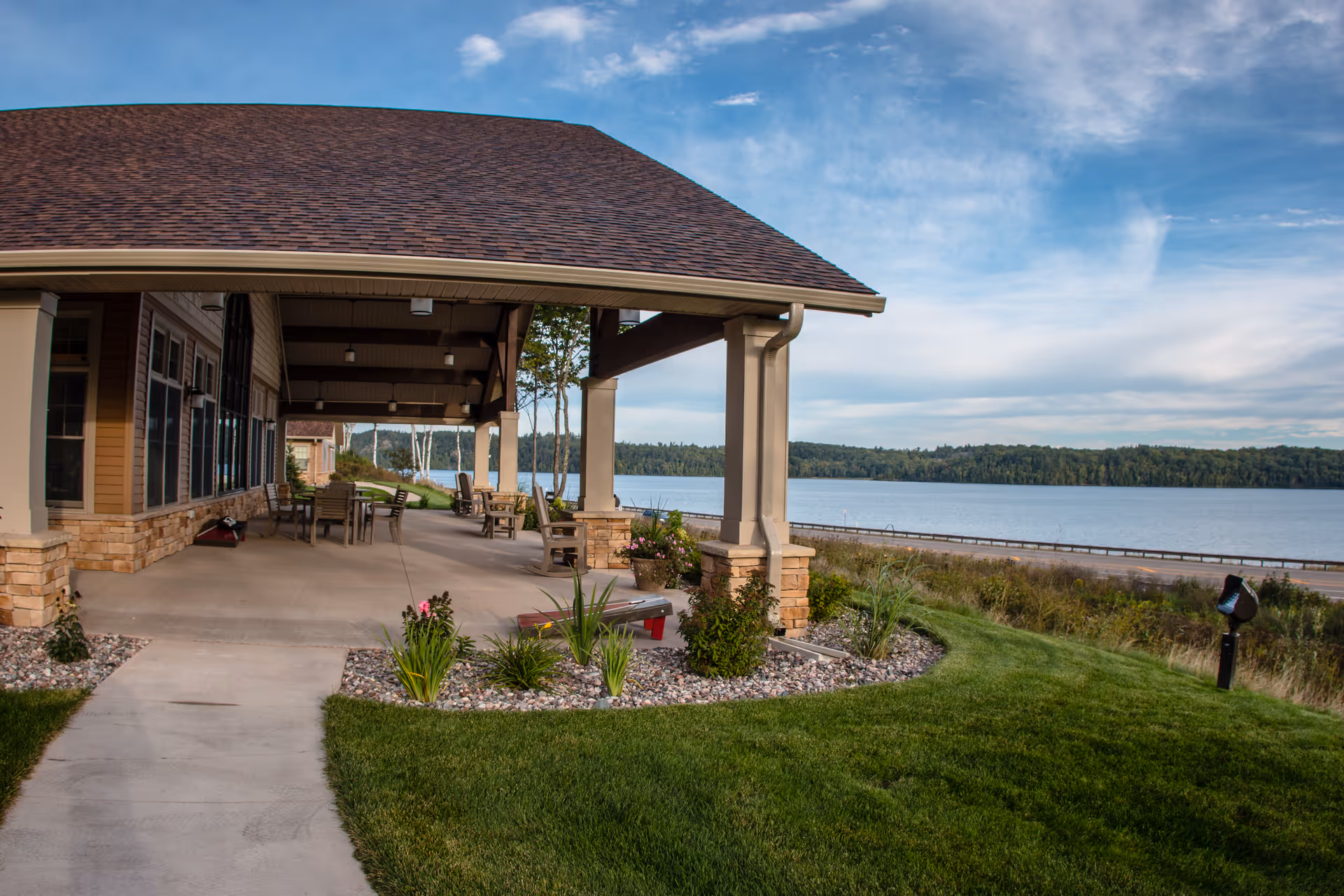 Covered outdoor patio area with tables and chairs next to a building, overlooking a large body of water with a tree-lined shore in the distance under a partly cloudy sky.