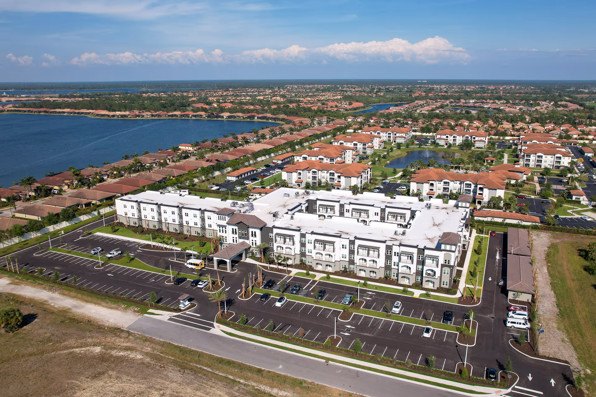 Aerial view of a multi-story senior living complex with parking lots, surrounding residential neighborhoods, and a nearby lake.