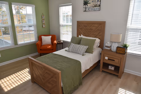 Sunny single-occupancy bedroom with a wooden bed dressed in green accents, a nightstand with a lamp and alarm clock, an orange armchair, and multiple windows.