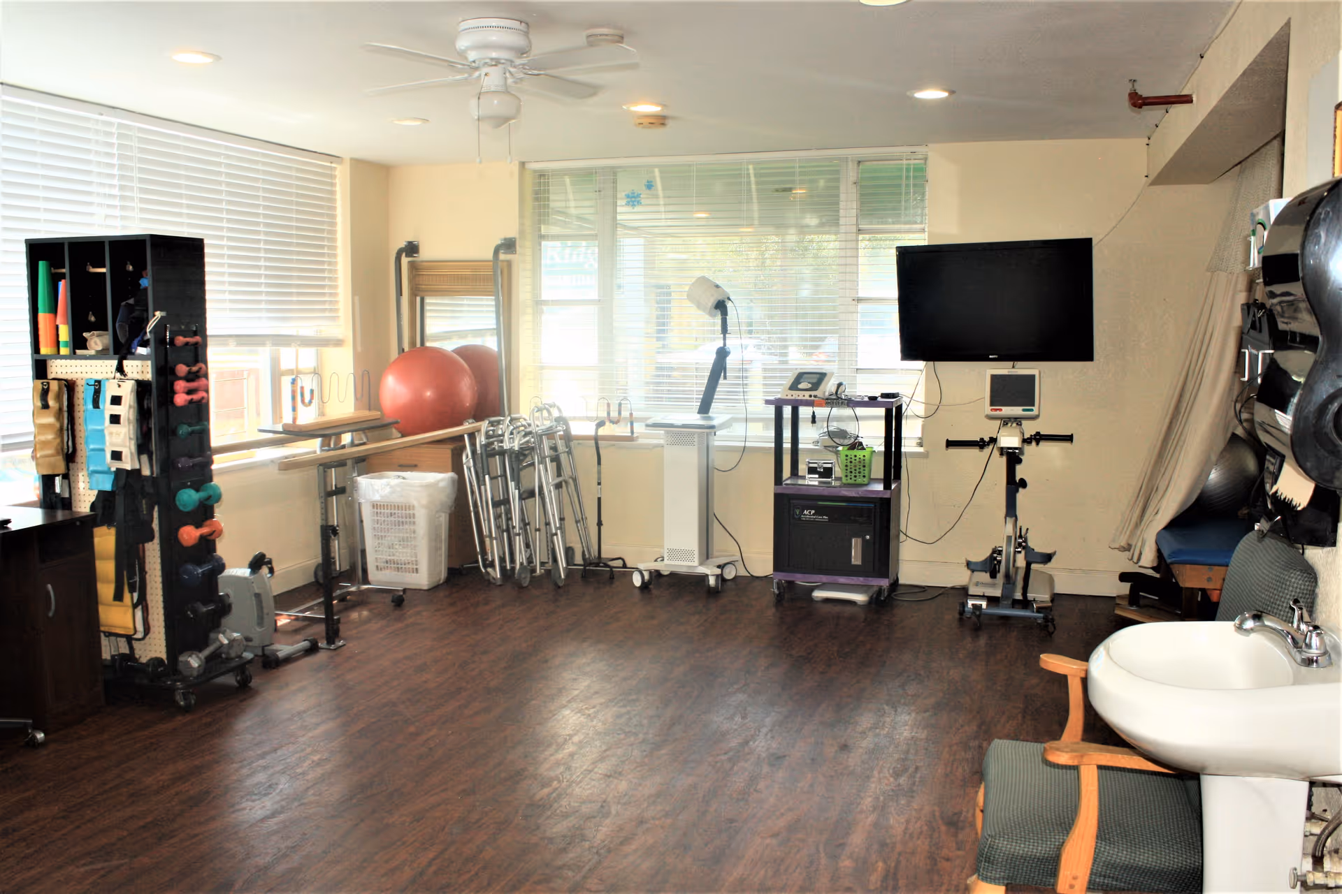 A rehabilitation or physical therapy room with exercise equipment including weights, resistance bands, walkers, a large red exercise ball, a stationary bike, and medical devices. The room has large windows with blinds, wood flooring, a ceiling fan, and a sink with a chair nearby.