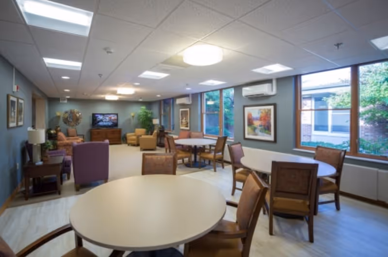 Communal dining and lounge area with round tables, chairs, armchairs, a TV and large windows.
