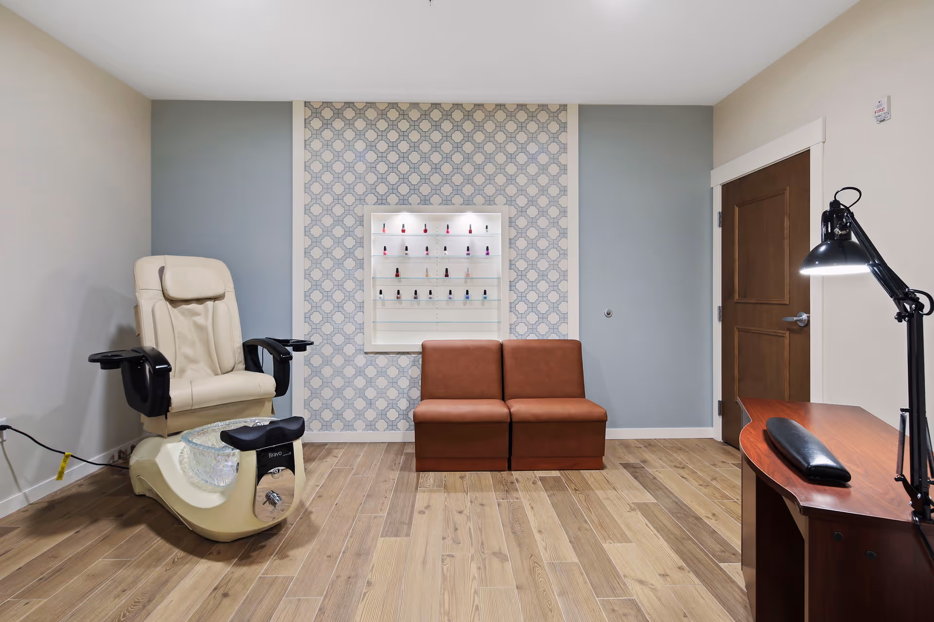 A nail salon room with a beige pedicure chair on the left, a small brown two-seat sofa in the center against a wall with patterned wallpaper, a display case with various nail polish bottles, and a wooden desk with a black adjustable lamp on the right.