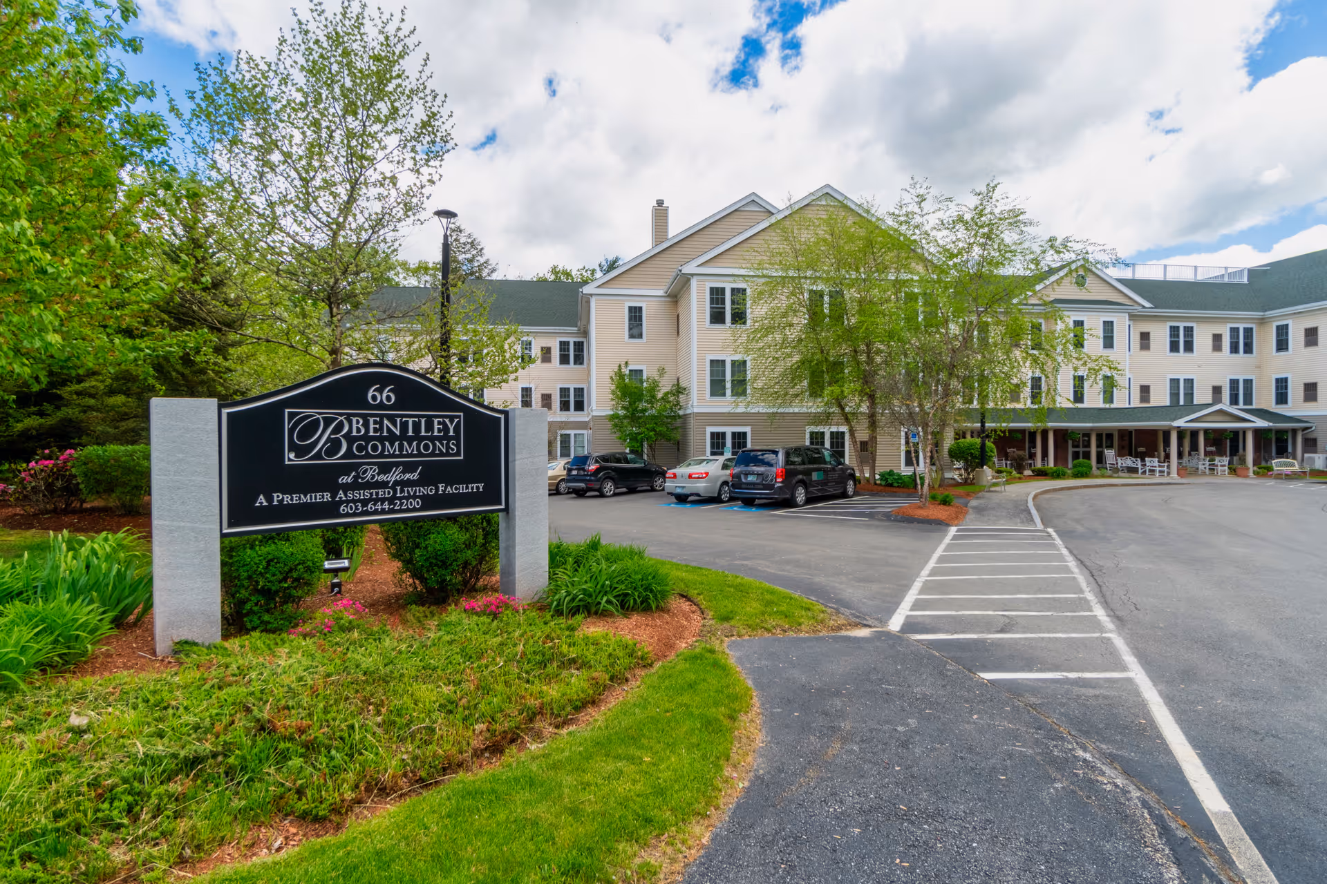 Exterior view of Bentley Commons at Bedford, a three-story assisted living facility with beige siding and green roof. The building is surrounded by trees and landscaping, with a parking lot in front. A large black and white sign near the entrance displays the facility name and contact information.