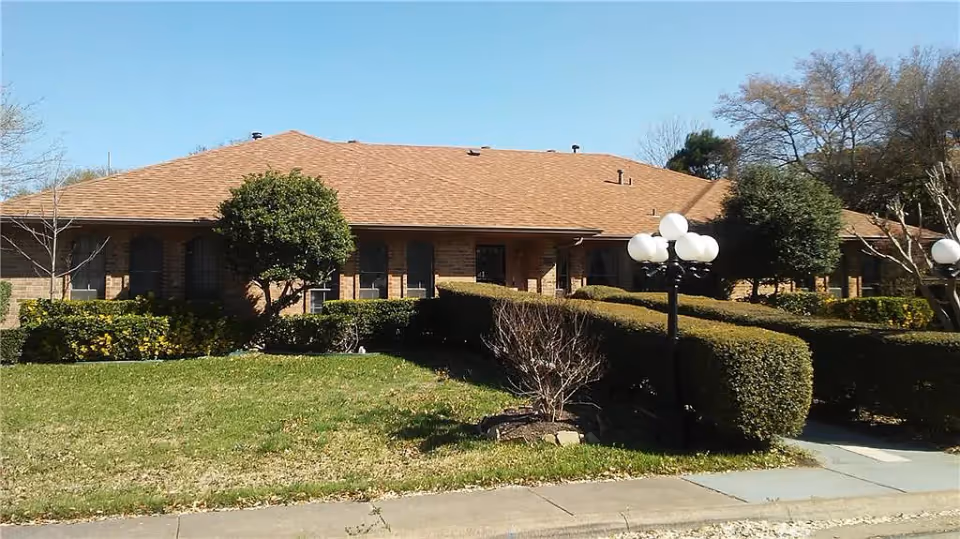 Single-story brick building with a brown shingle roof, surrounded by trimmed bushes, trees, and a green lawn under a clear blue sky. A sidewalk runs in front of the building, and there are black lamp posts with multiple white globe lights near the entrance.