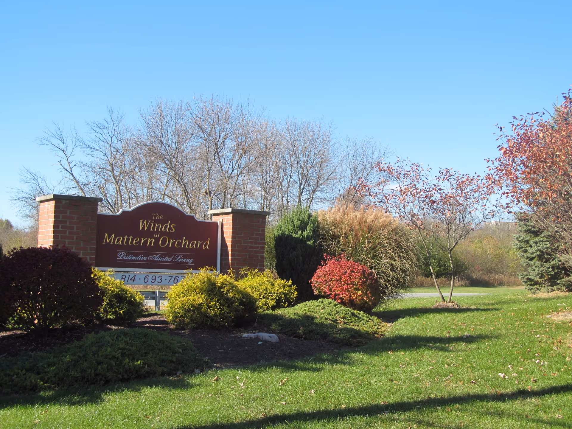 Outdoor view of a landscaped area with green grass, bushes, and trees with autumn foliage surrounding a brick sign that reads 'The Winds at Mattern Orchard Distinctive Assisted Living'. The sky is clear and blue.