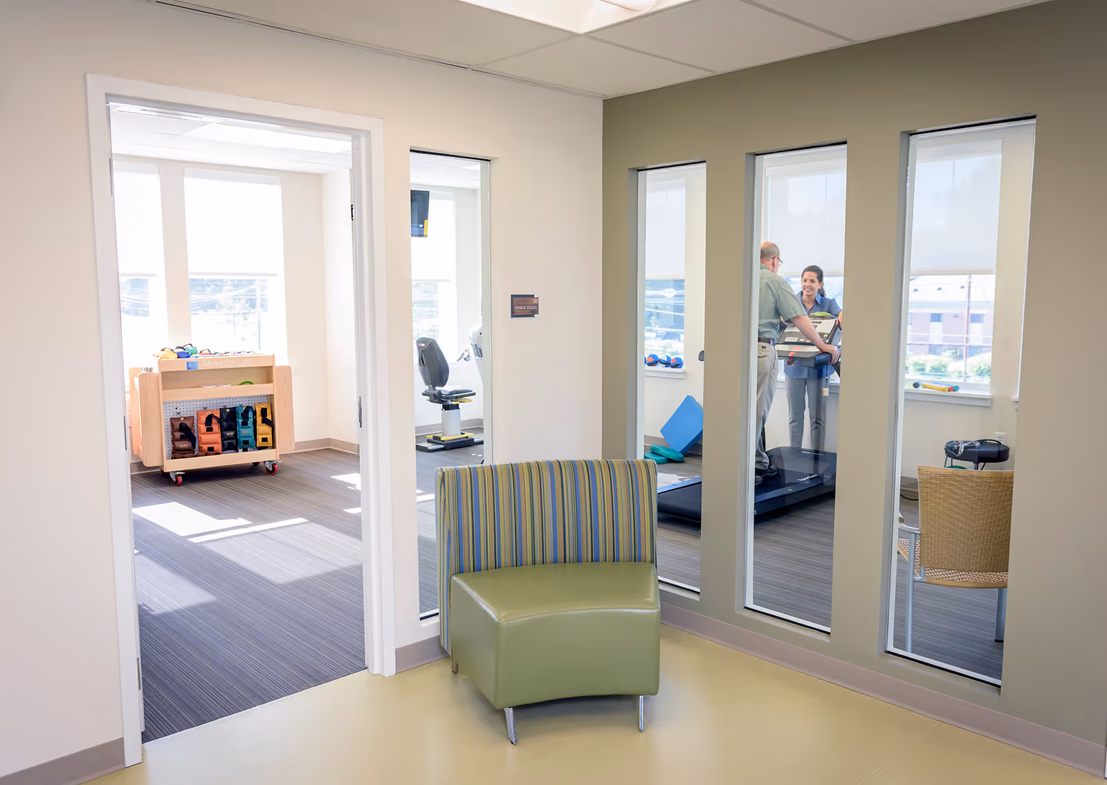 View of a fitness or rehabilitation room through a glass wall, showing a man walking on a treadmill assisted by a staff member. In the foreground, there is a green and striped chair in a small waiting or hallway area. The adjacent room contains exercise equipment and weights on a rack.